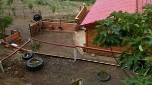 Una casa de madera rústica con techo rojo, rodeada por un patio cercado con un sendero de tierra, y montañas verdes exuberantes al fondo.