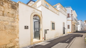 A street with a white building featuring an arched entrance, a small window, and a mailbox. - 3396 Leucadense - Grotta 1 by Barbarhouse (Castrignano del Capo)