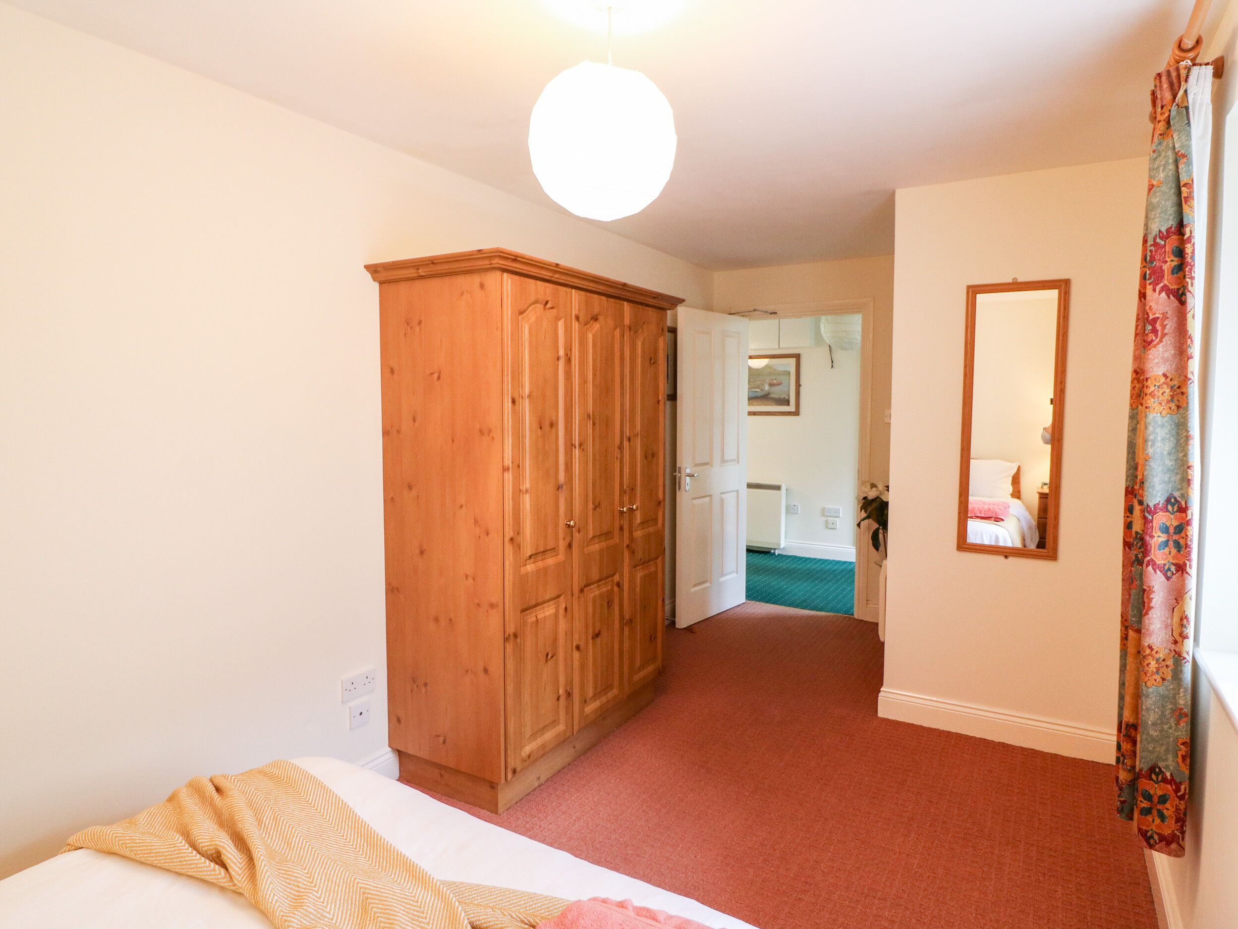 Un dormitorio con un armario de madera, una cama con manta a rayas, un espejo y una puerta que da a otra habitación.