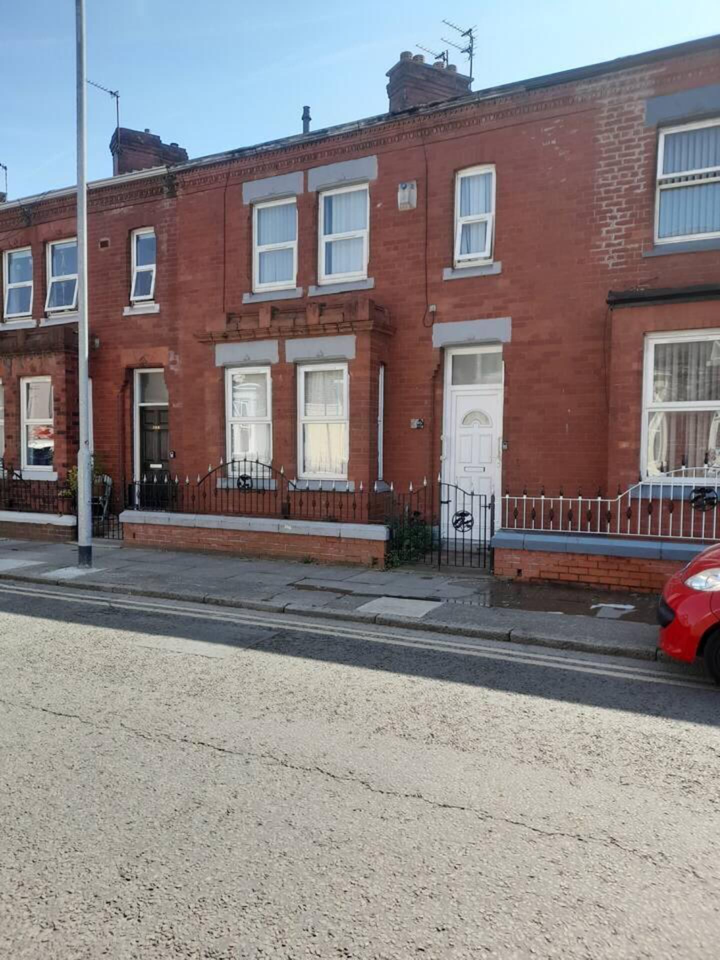 A row of red brick terraced houses with white doors and windows, a metal fence, and a red car parked on the street.