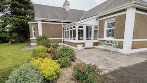 A house with a conservatory, a garden with various plants and flowers, and a bench outside. - CHURCHFIELD HOUSE, pet friendly in Crossmolina, County Mayo (Ballina)