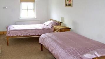 A room with two single beds, a window with blinds, a bedside table with a lamp, and a framed picture on the wall.