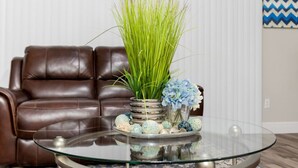 A glass coffee table with a vase of green grass and blue flowers, a brown leather couch, and a blue zigzag patterned wall. - 3BR Townhouse Right in Front of Epic Universe! (Orlando)