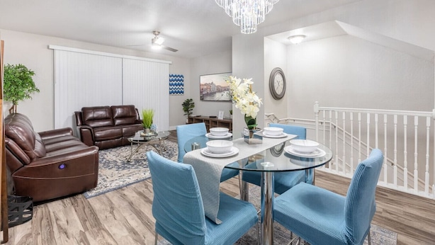 A modern living room with a brown leather sofa, a glass coffee table, and blue chairs. There is a dining area with a glass table and white plates. A chandelier hangs from the ceiling, and a clock is on the wall. - 3BR Townhouse Right in Front of Epic Universe! (Orlando)