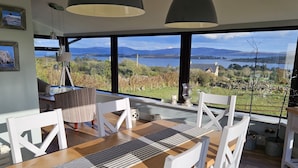 En matplats med ett träbord och vita stolar, stora fönster med utsikt över en sjö och berg.