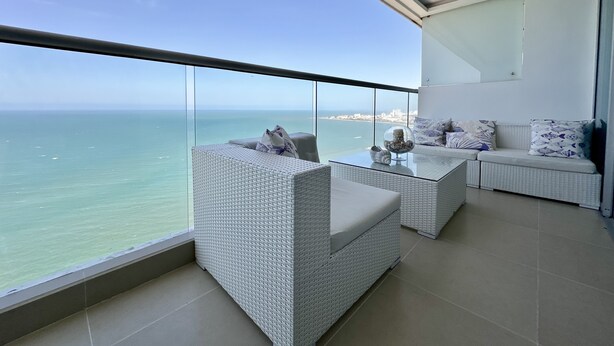 Ein Balkon mit Glastrennwand, weißen Korbmöbeln und Blick auf das Meer und die Stadtlandschaft.
