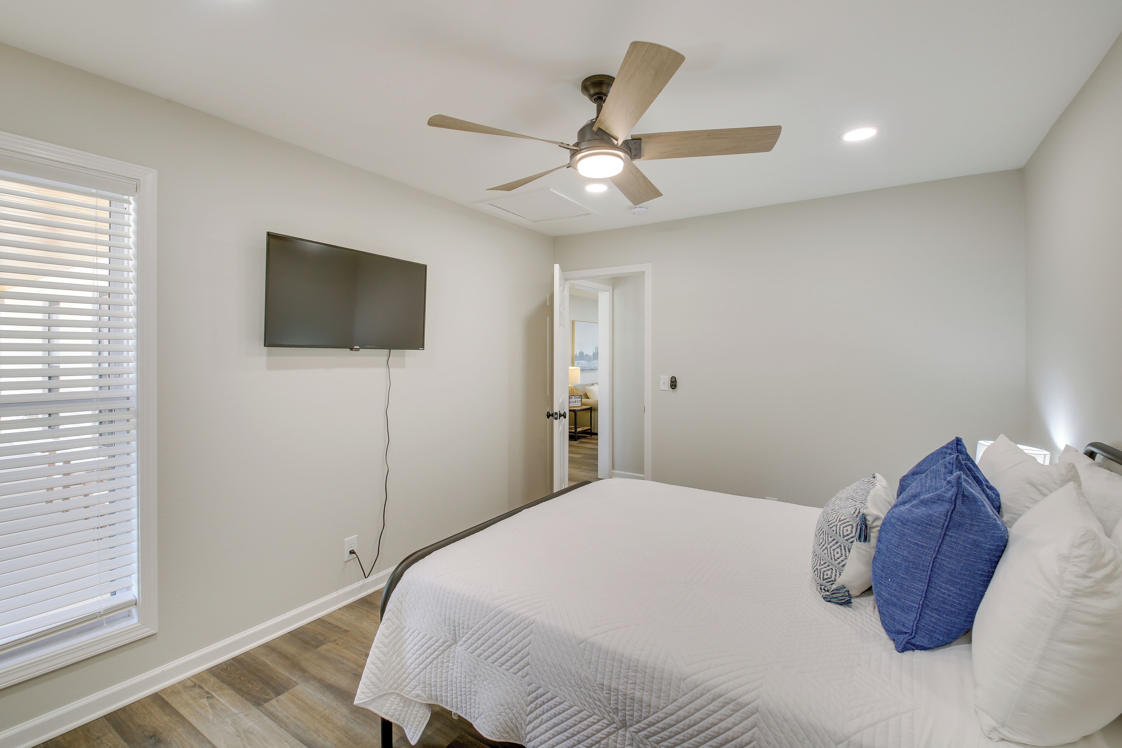 Un dormitorio con cama, ventilador de techo, televisor montado en la pared y una puerta que da a otra habitación.