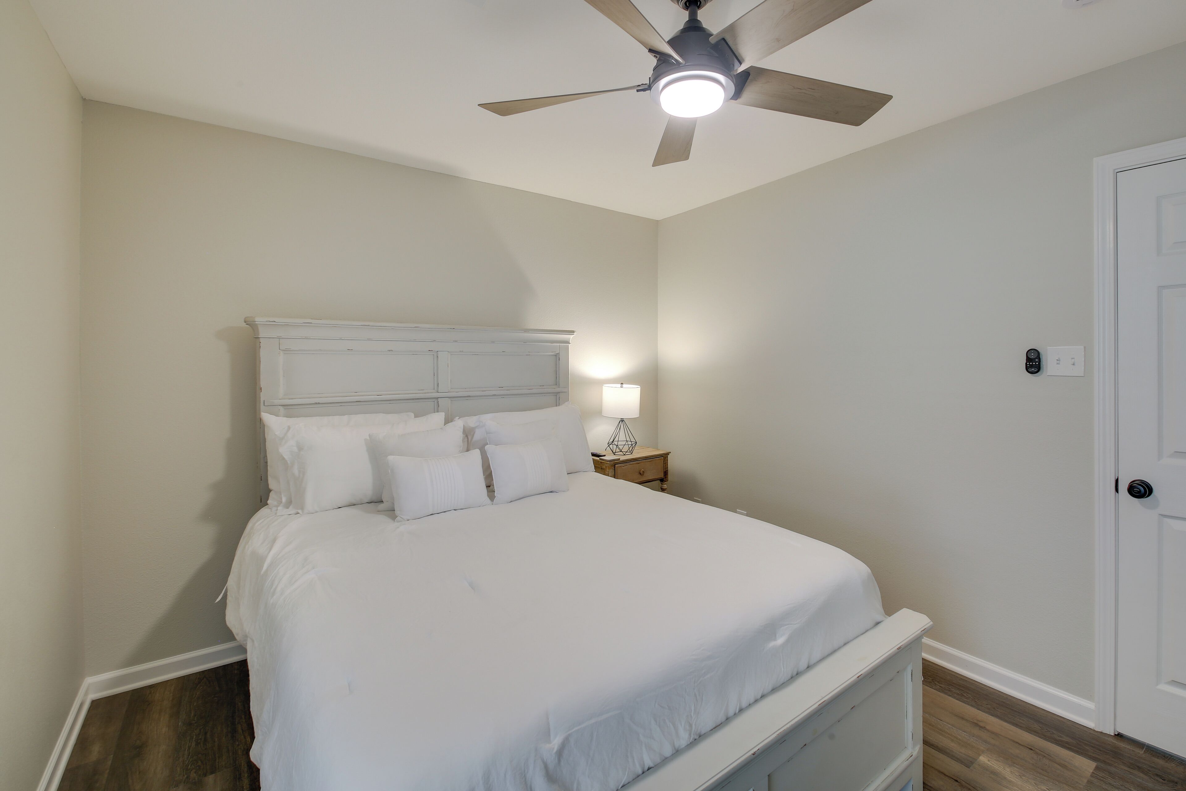 A neatly made bed in a room with a ceiling fan, a bedside lamp, and a door.
