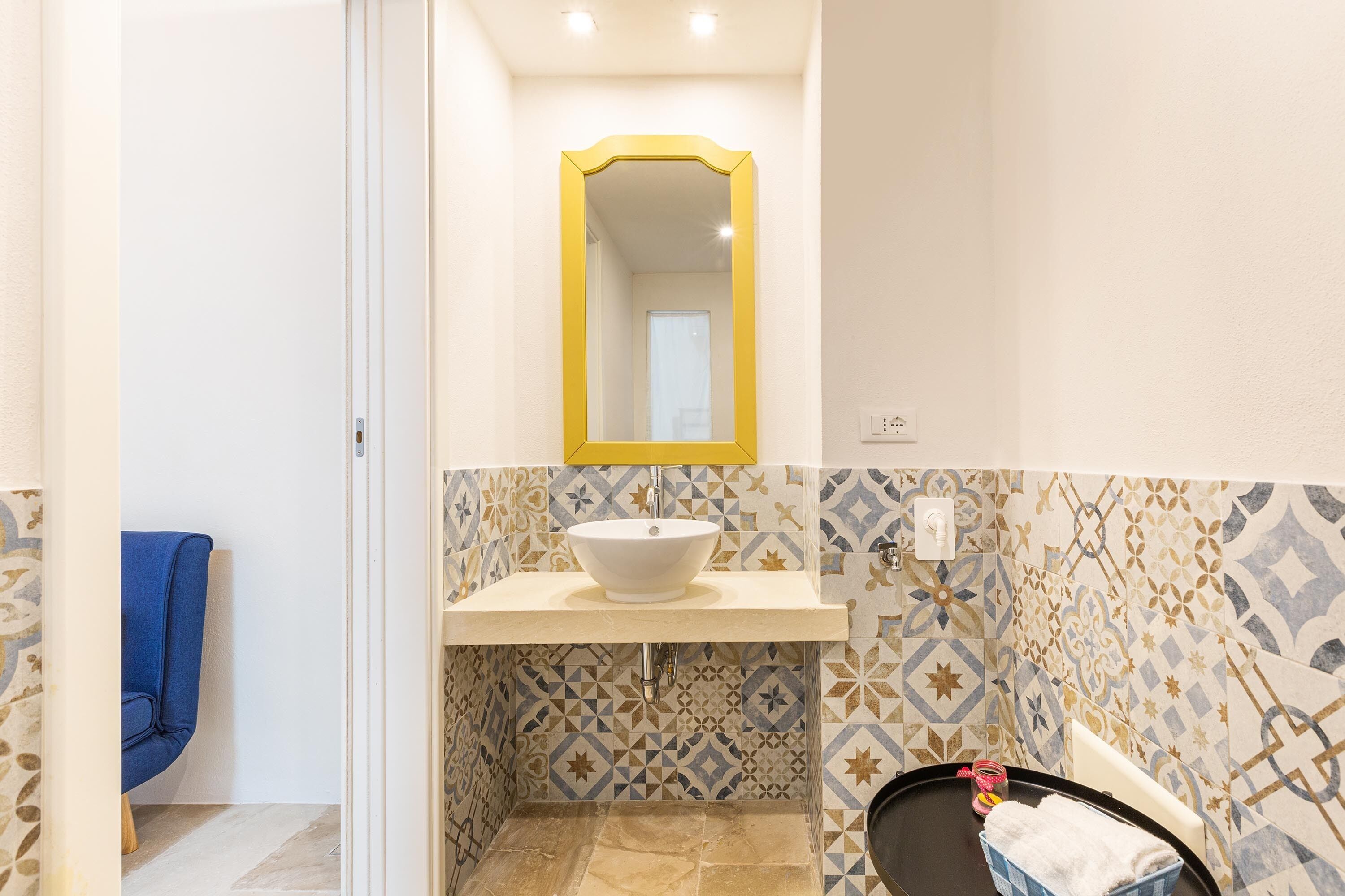 A bathroom with a white sink, a yellow-framed mirror, patterned tiles, and a blue chair.