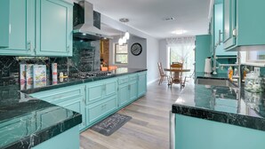 Een keuken met smaragdgroene kastjes, zwarte marmeren aanrechtbladen en een eetgedeelte met een tafel en stoelen.