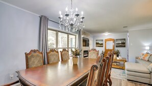 Een eetkamer met een grote houten tafel, zes gestoffeerde stoelen, een kroonluchter en een groot raam met gordijnen.