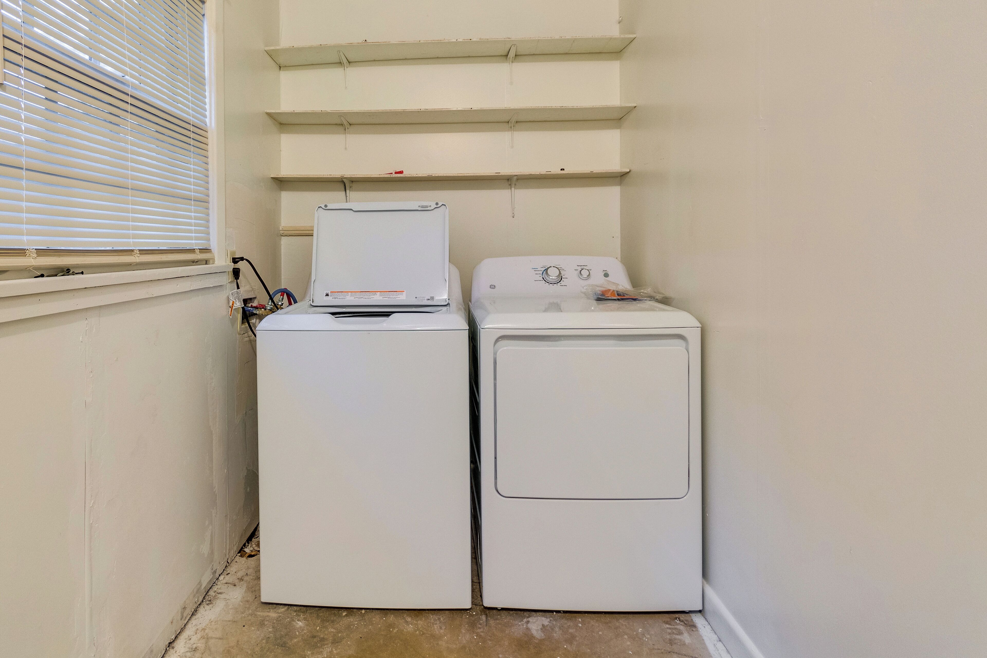 A laundry room with two stacked washing machines and a window with blinds.