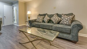 Una sala de estar con una mesa de centro de vidrio, un sofá gris con cojines estampados, una lámpara y suelo de madera.