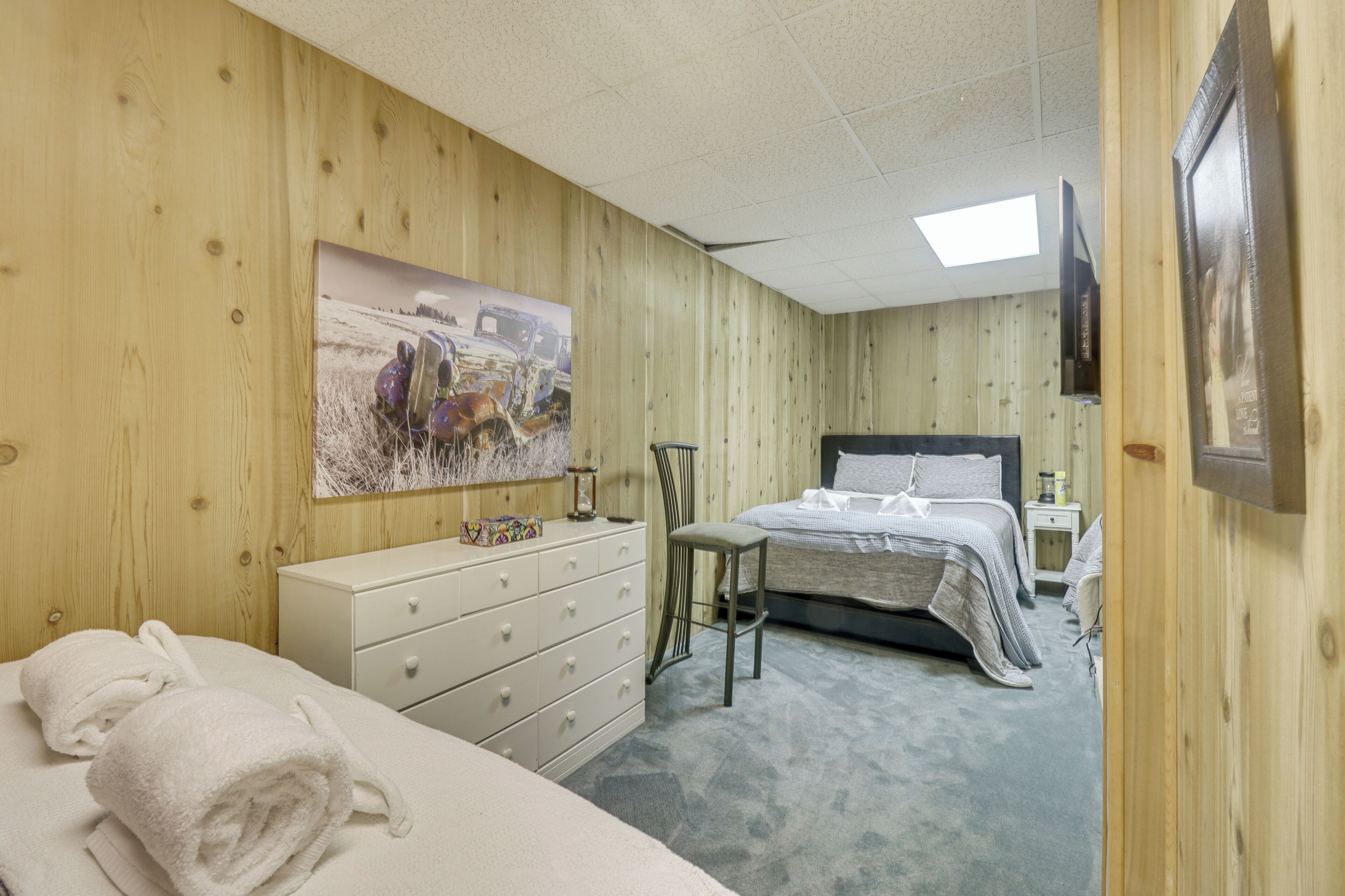 Een kamer met houten wanden, een bed met een grijs-wit gestreepte dekbedovertrek, een witte dressoir en een schilderij van een veld met een vrachtwagen en paarden.