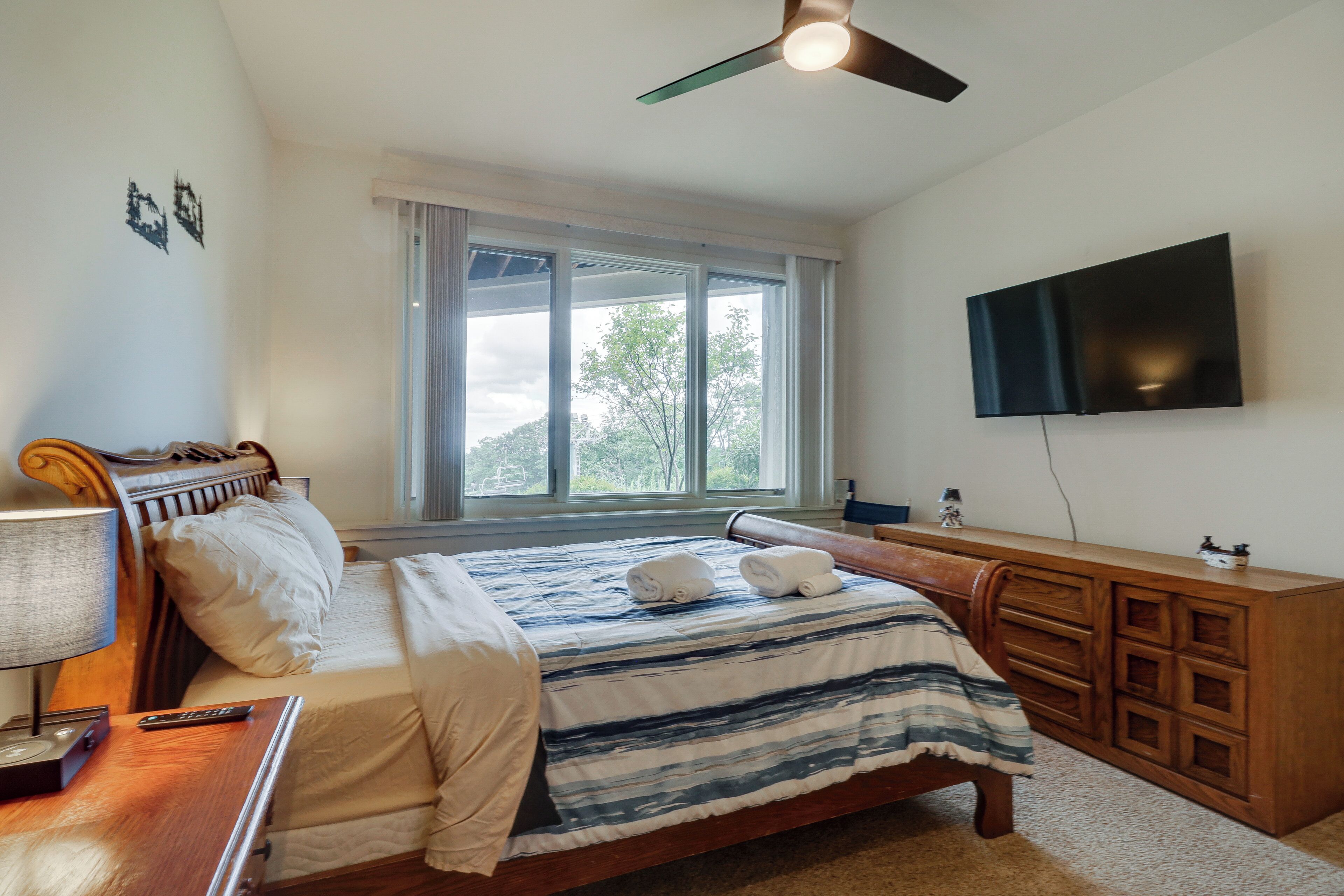 A bedroom with a bed, a wooden dresser, a television mounted on the wall, and a window with curtains.