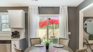 Un coin repas avec une table ronde dressée pour quatre personnes, une machine à café et une vue sur un jardin avec un parasol rouge.