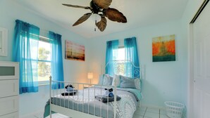 Ein Schlafzimmer mit Bett, Vorhängen, Deckenventilator, Fenster, Wandbild und Wäschekorb.