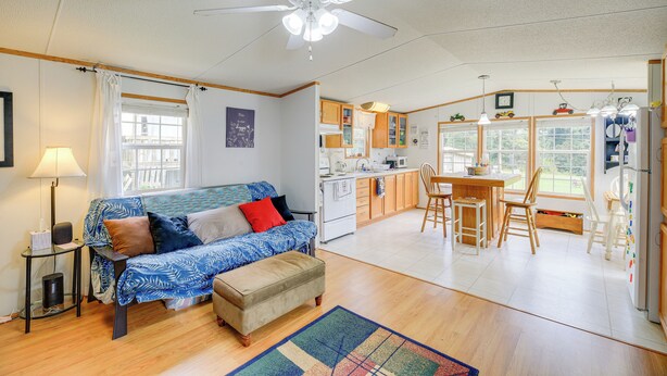 A cozy living room with a sofa, ottoman, and a ceiling fan. A kitchen with wooden cabinets and a dining area is visible in the background. - Creekfront Max Meadows Mobile Home With Deck! (Max Meadows)