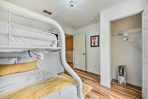 Un lit superposé avec une literie blanche et une couverture jaune, un plancher en bois, une porte blanche et un tableau encadré au mur.