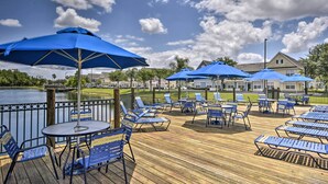 Terraza de madera con sillas y sombrillas azules, con vistas a un lago y una fila de casas.