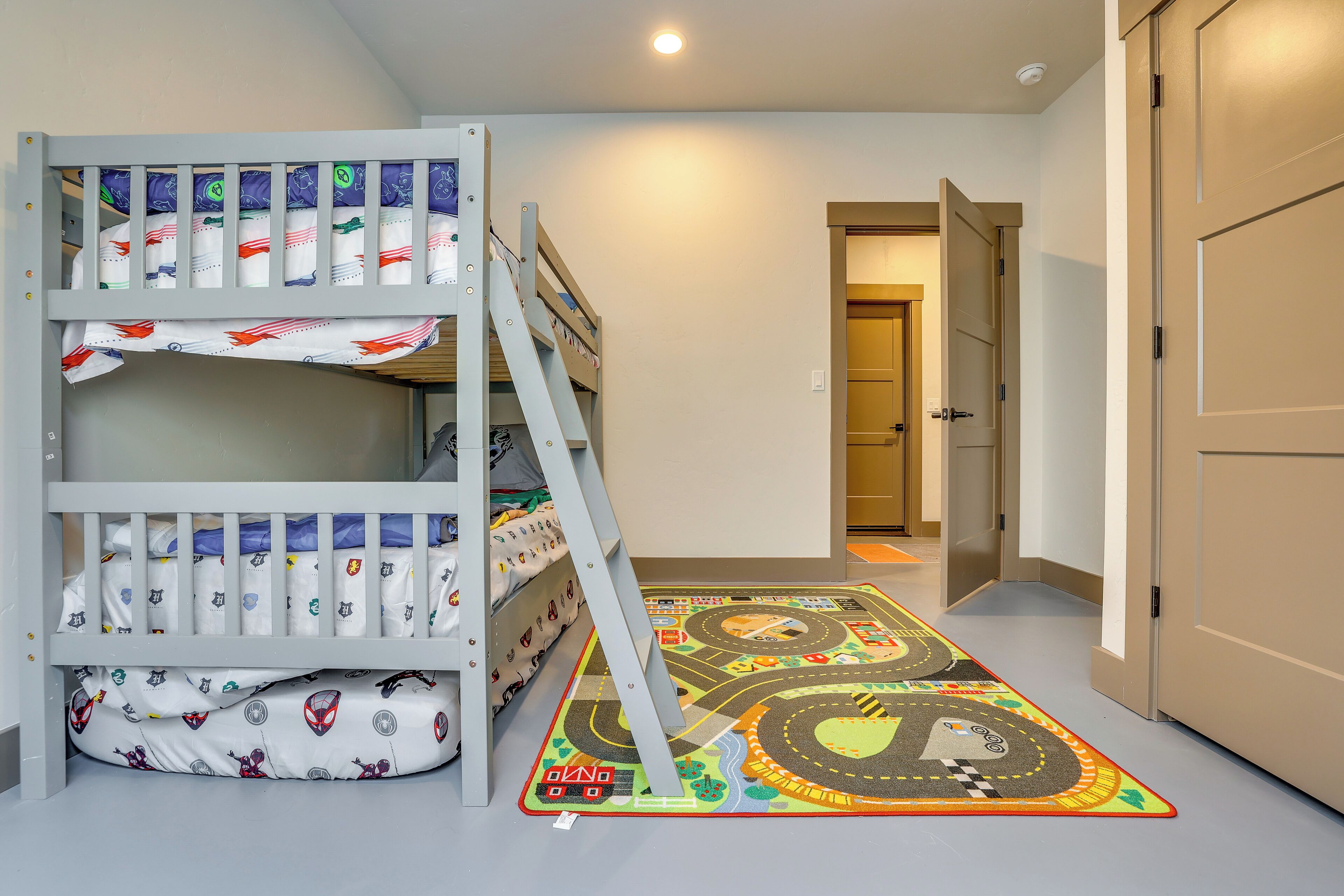 Une chambre d’enfant avec des lits superposés, un tapis de jeu coloré et un couloir menant à une porte.