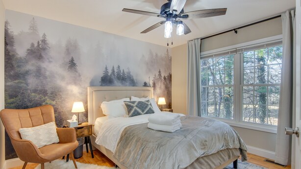 A bedroom with a large bed, a chair, a nightstand with a lamp, a ceiling fan, and a window with a view of trees. - Asheville Retreat ~ 6 Mi to Biltmore & Downtown! (Asheville)