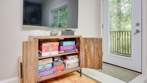 A room with a TV mounted on the wall, a wooden shelf with various board games, and a door leading to a balcony with a view of trees. - Asheville Retreat ~ 6 Mi to Biltmore & Downtown! (Asheville)
