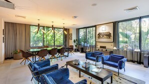 A modern living room with blue sofas, a dining area with a table and chairs, large windows with a view of greenery, and a kitchen area with a stove and cabinets. - High-end Miami Condo w/ City & Bay Views!!! (Miami)