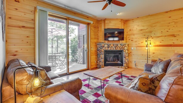 A cozy living room with a stone fireplace, a ceiling fan, and a large sliding glass door leading to an outdoor patio. - Walk to Main Street: Grand Lake Condo w/ Patio (Grand Lake)
