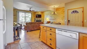 A kitchen with wooden cabinets, a dishwasher, a dining area with a table and chairs, and a TV mounted above the fireplace. - Walk to Main Street: Grand Lake Condo w/ Patio (Grand Lake)