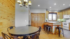 A kitchen with wooden cabinets, a central island with stools, a dining table with chairs, and a chandelier. - Walk to Main Street: Grand Lake Condo w/ Patio (Grand Lake)