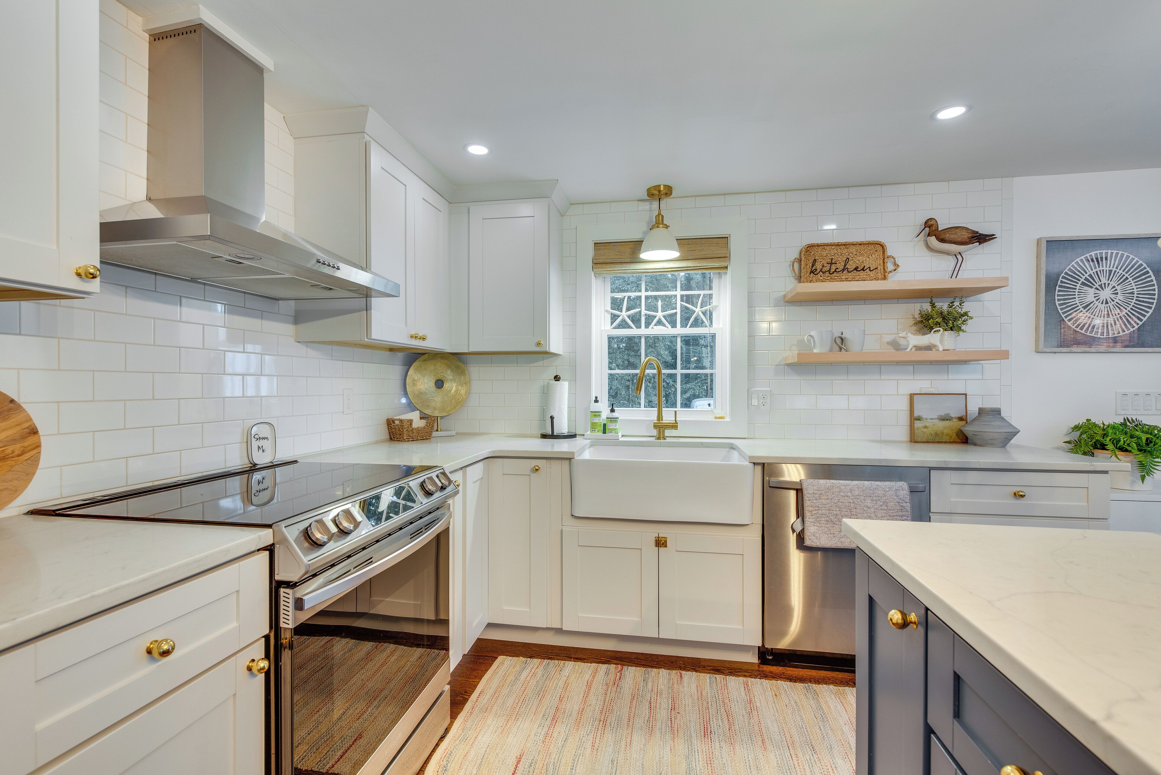 A kitchen with white cabinets, a stainless steel range hood, a built-in oven, a dishwasher, and a window with a view of the outdoors.