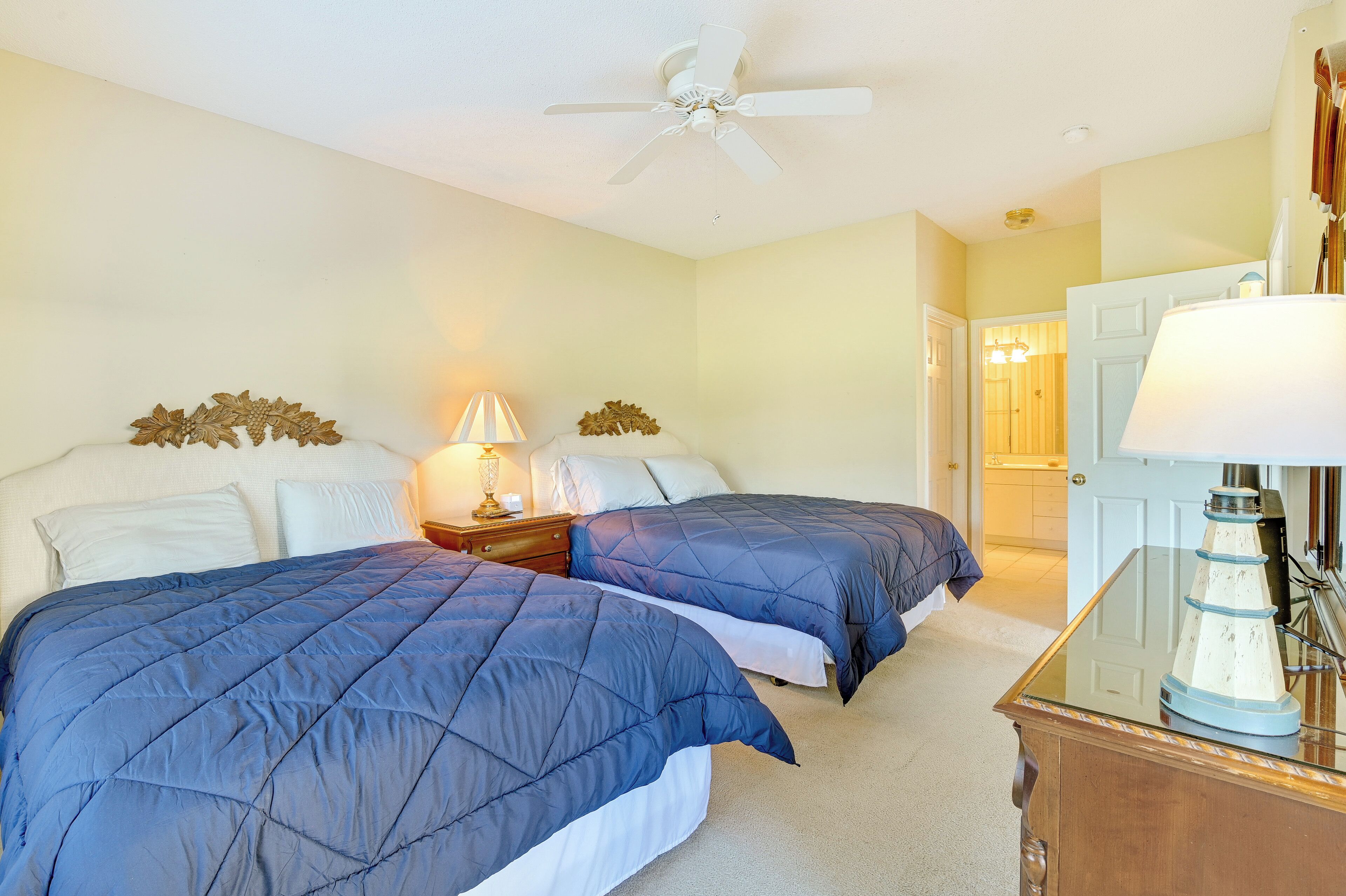 A bedroom with two beds, a ceiling fan, a nightstand with a lamp, and a dresser with a decorative item.