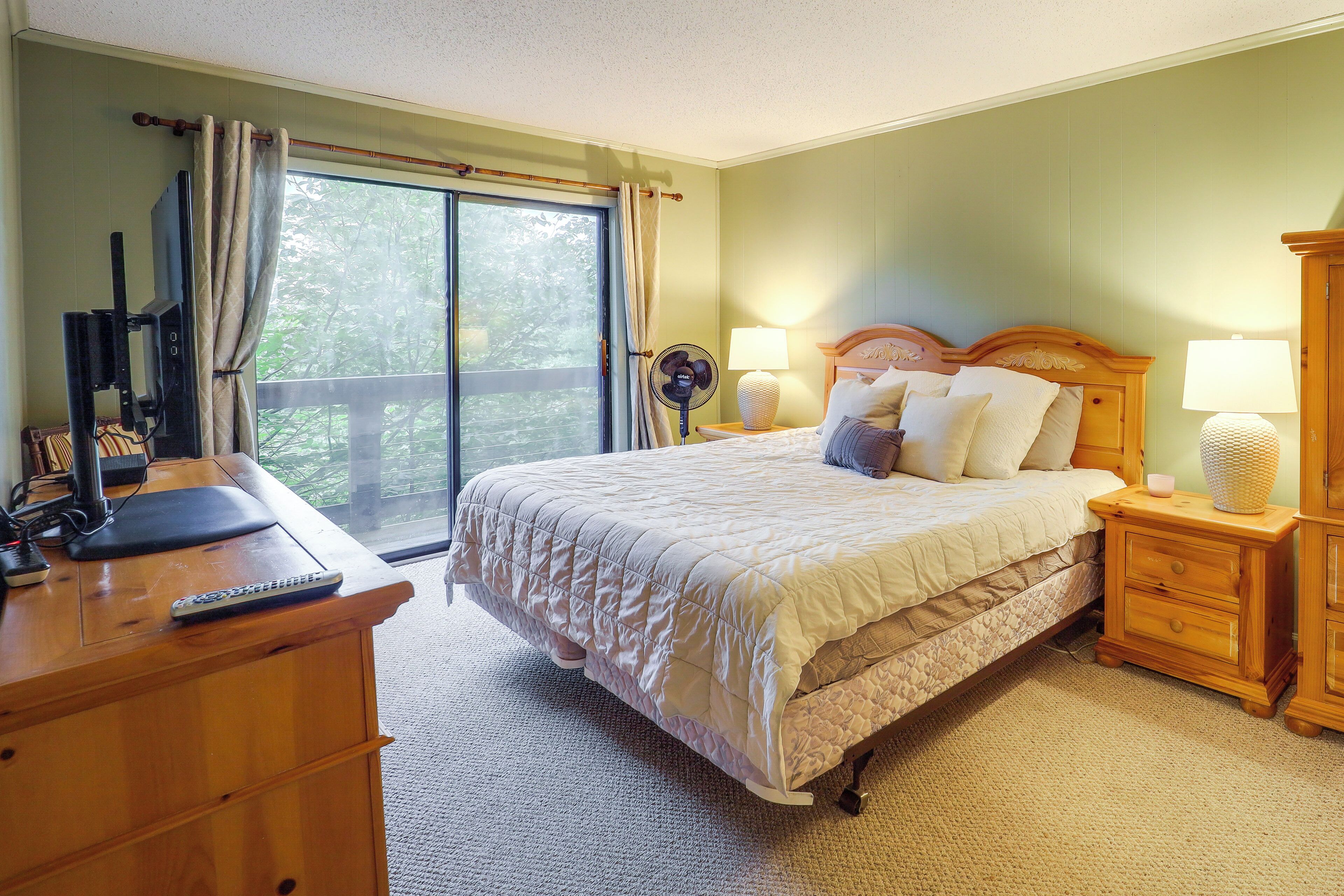 A bedroom with a bed, bedside lamps, a wooden nightstand, a desk with a computer, and a window with curtains.