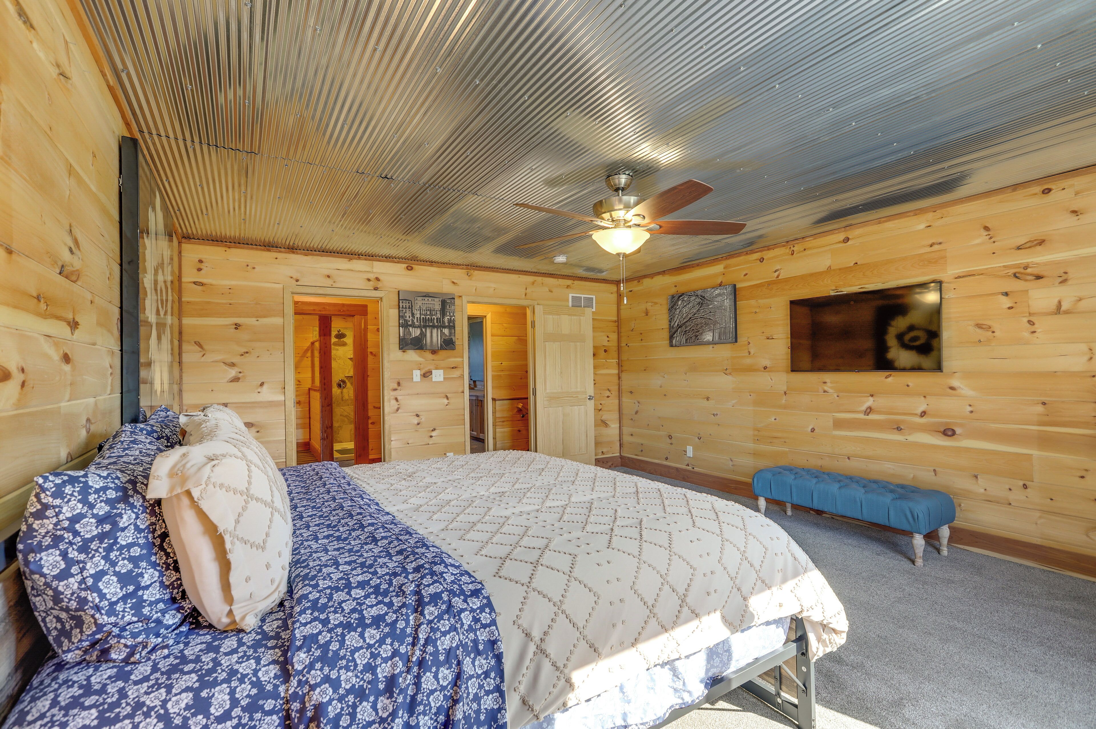 Quarto de cabana de madeira com uma cama, um banco, um ventilador de teto e uma porta que dá para outro cômodo.