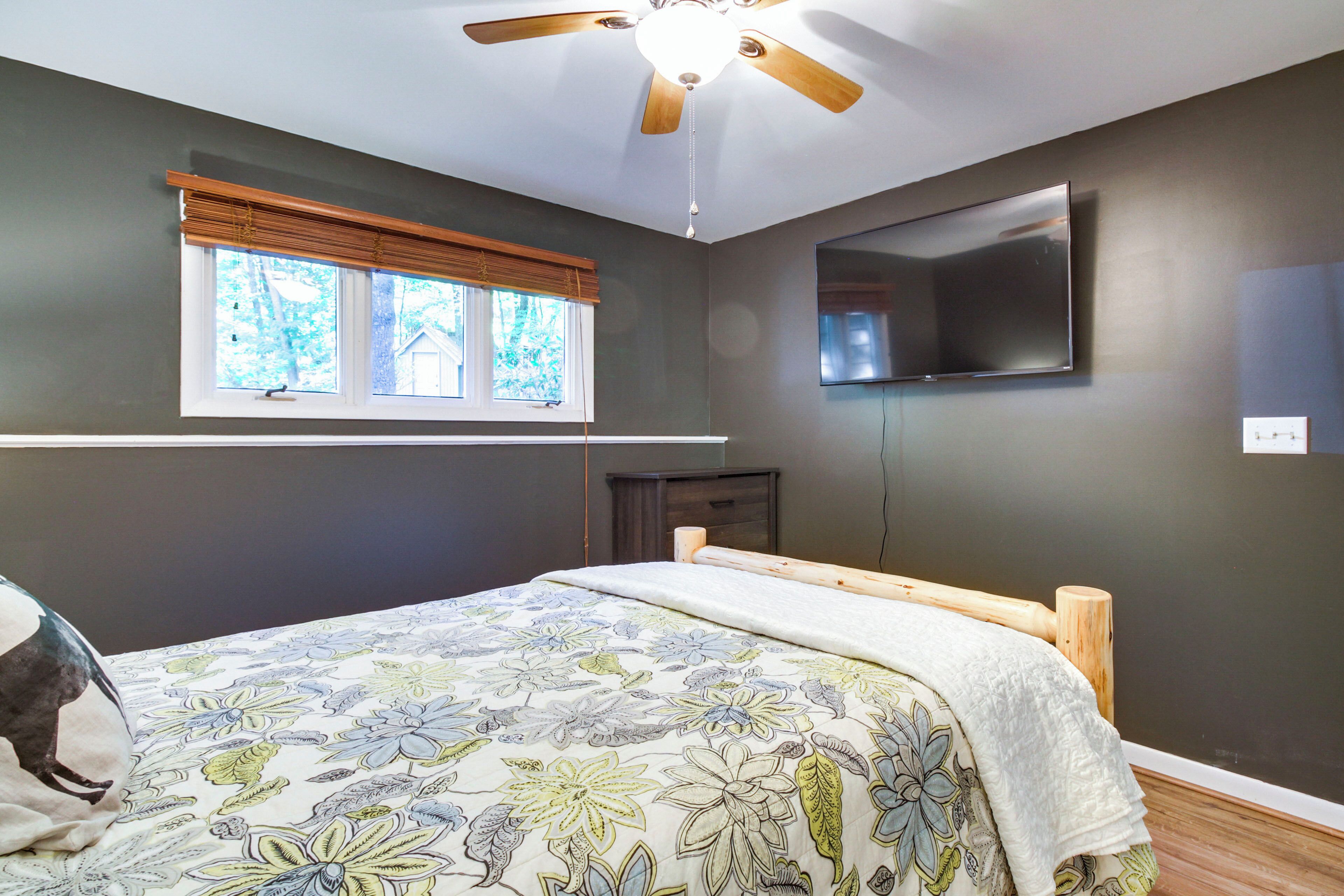 Un dormitorio con cama, ventilador de techo, ventana con persianas, televisor de pantalla plana y cabecero de madera.