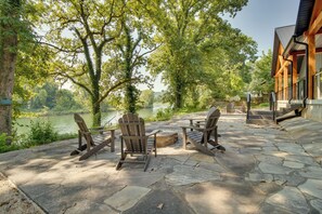 Un espace extérieur aménagé avec des chaises Adirondack donnant sur une rivière, un sol en pierre et un bâtiment en bois doté d’un porche.