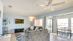 A spacious living area with a grey sofa, a dining table with chairs, a ceiling fan, and a large window providing natural light. - Bright Destin Condo w/ Community Pool! (Destin)