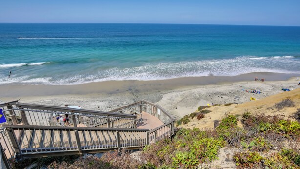 Una playa con una escalera de madera que baja a la orilla, arena y olas del océano.