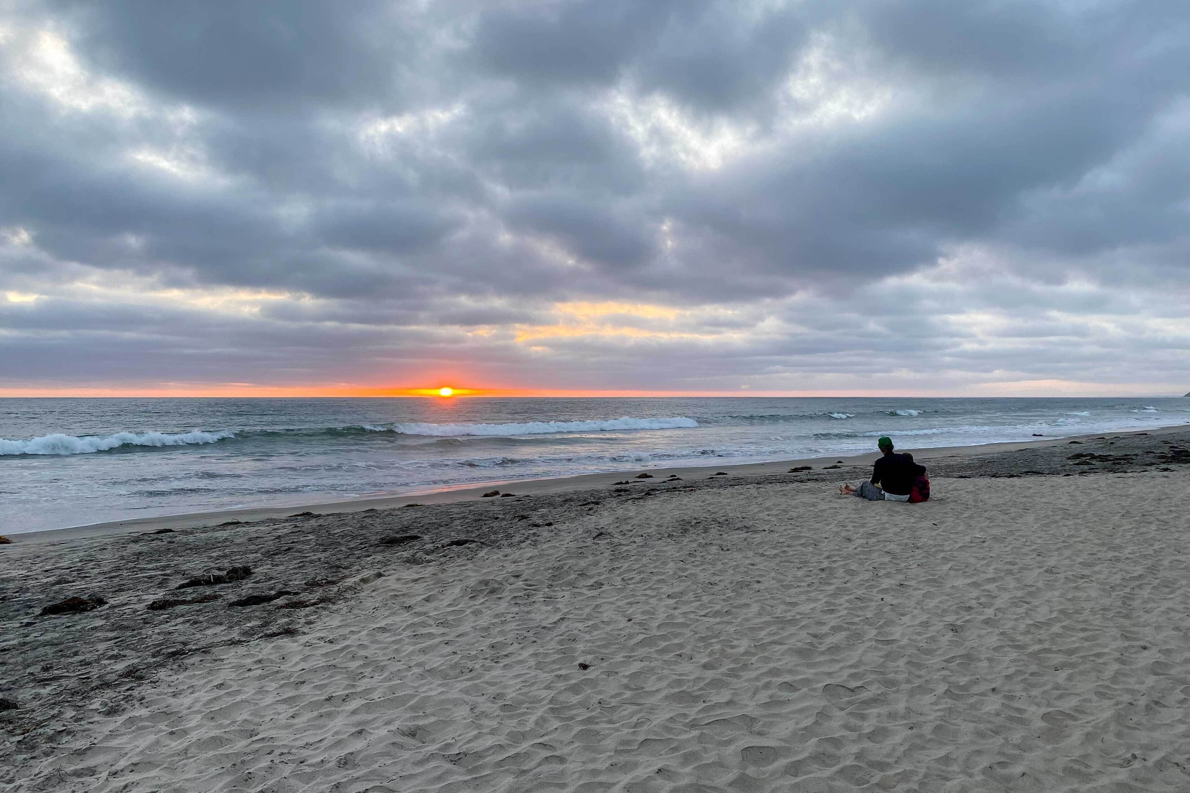A beach with a person sitting on the sand, facing the ocean during sunset.