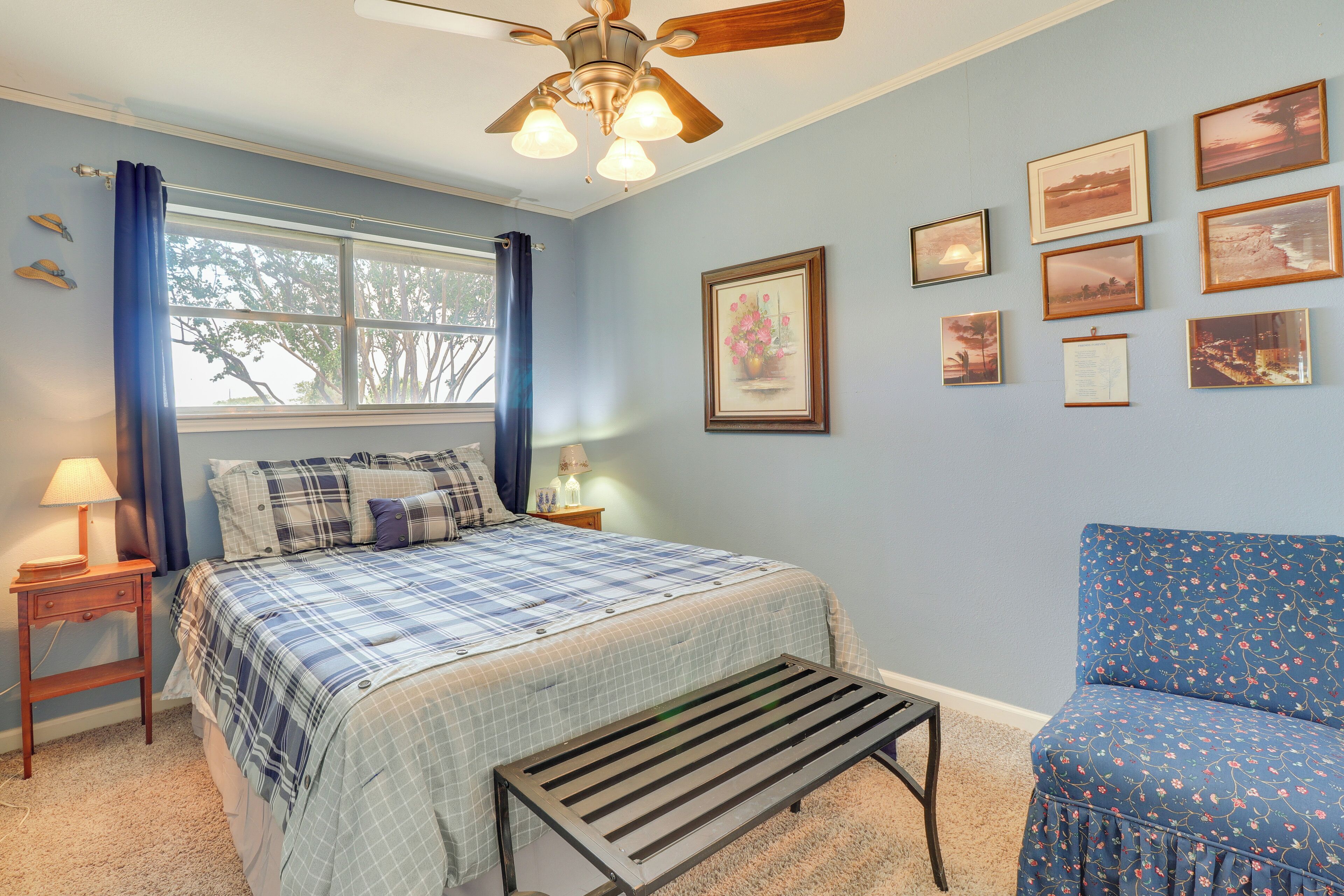 A bedroom with a bed, a chair, a ceiling fan, a window with curtains, and framed pictures on the wall.