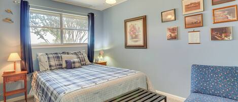 A bedroom with a bed, a chair, a ceiling fan, a window with curtains, and framed pictures on the wall.