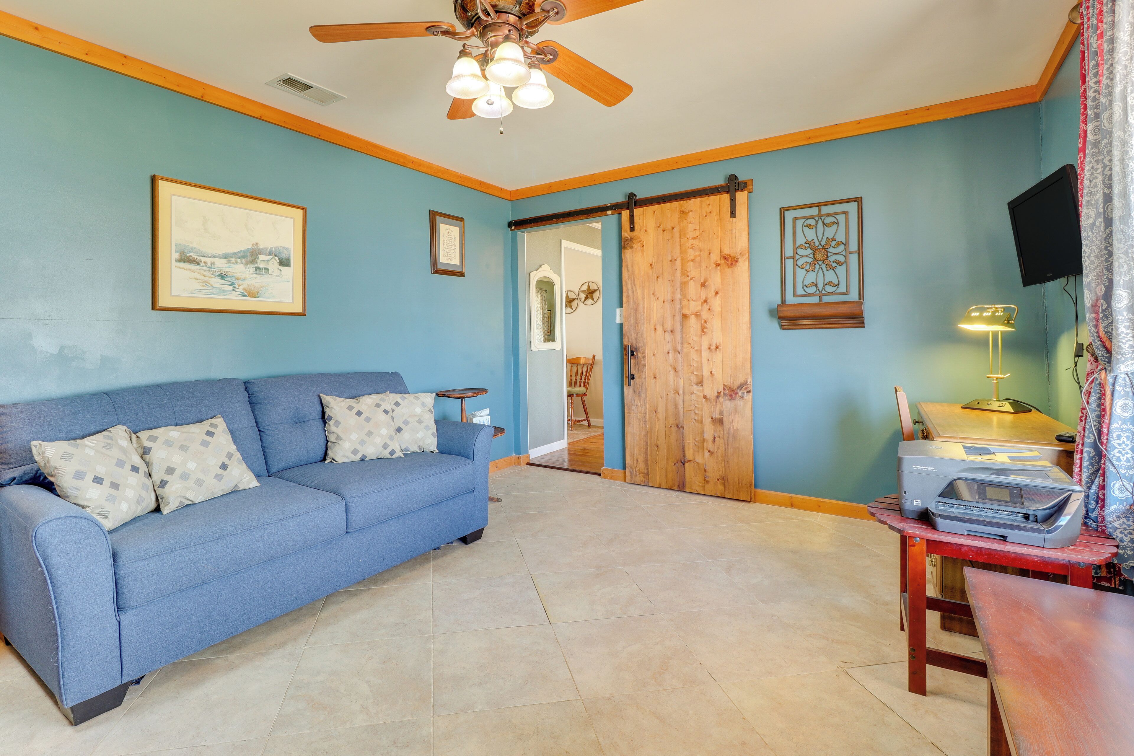 A living room with a blue sofa, a ceiling fan, a sliding door, and a table with a lamp and printer.