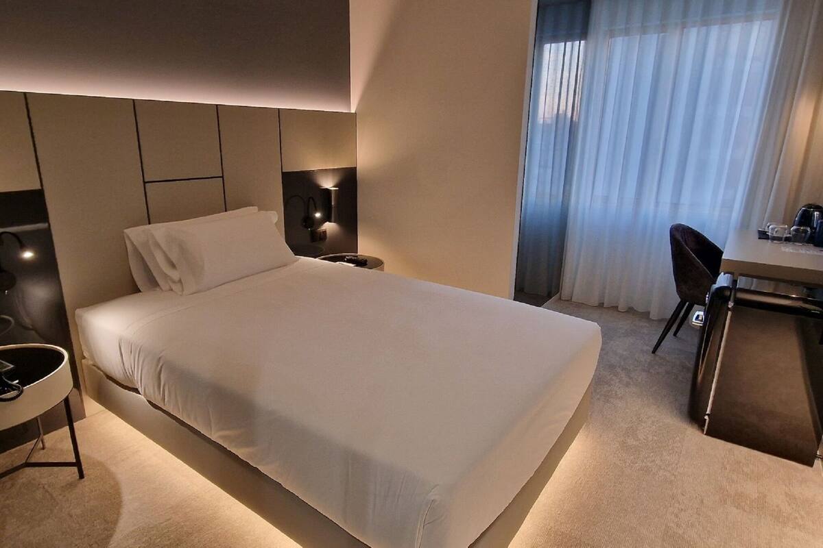 Una habitación de hotel moderna con una cama bien hecha, un escritorio con una silla, una mesita redonda y una lámpara de pared.