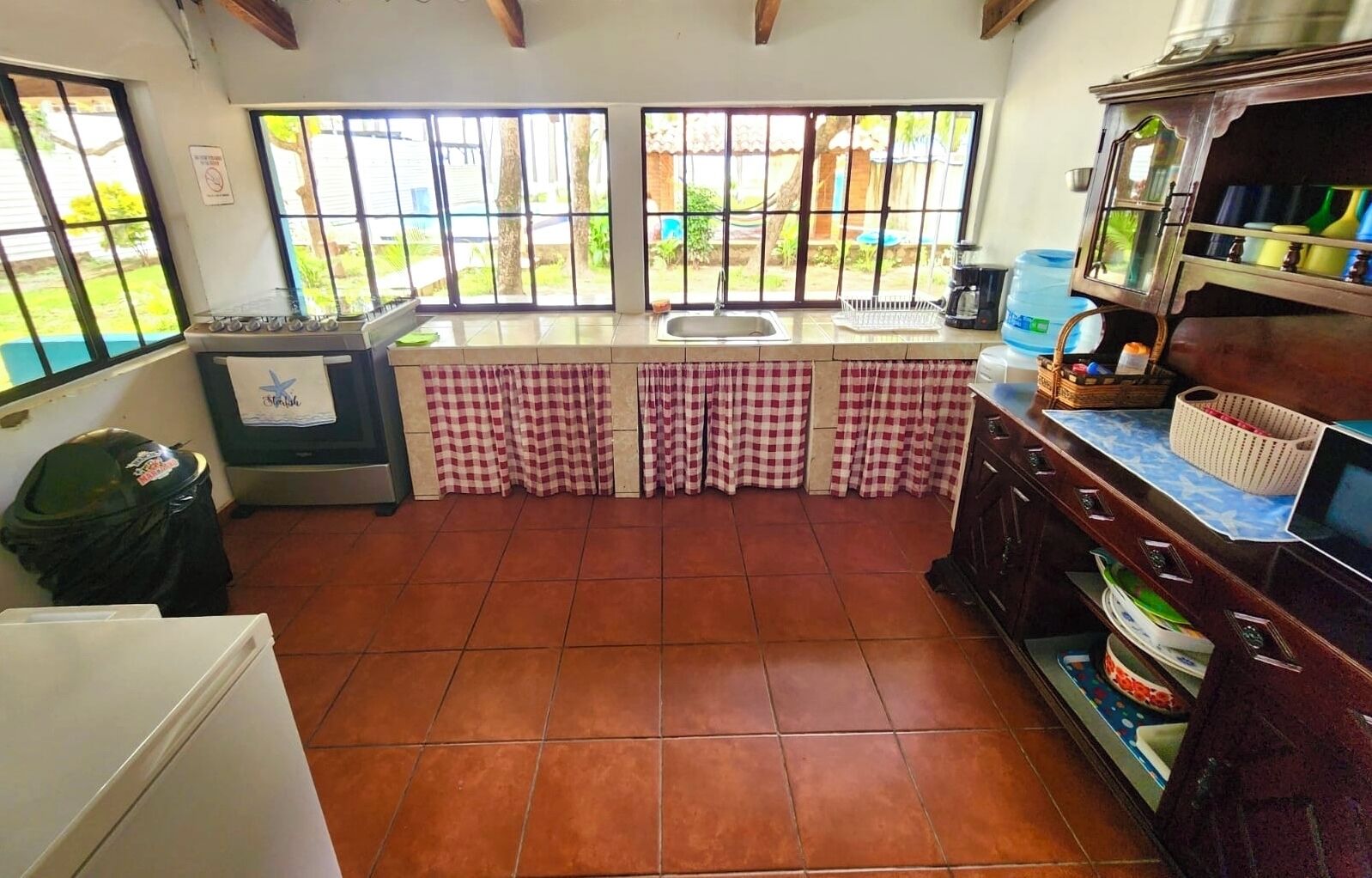 A kitchen with a tiled floor, a sink, a stove, a trash bin, and a window with a view of greenery.