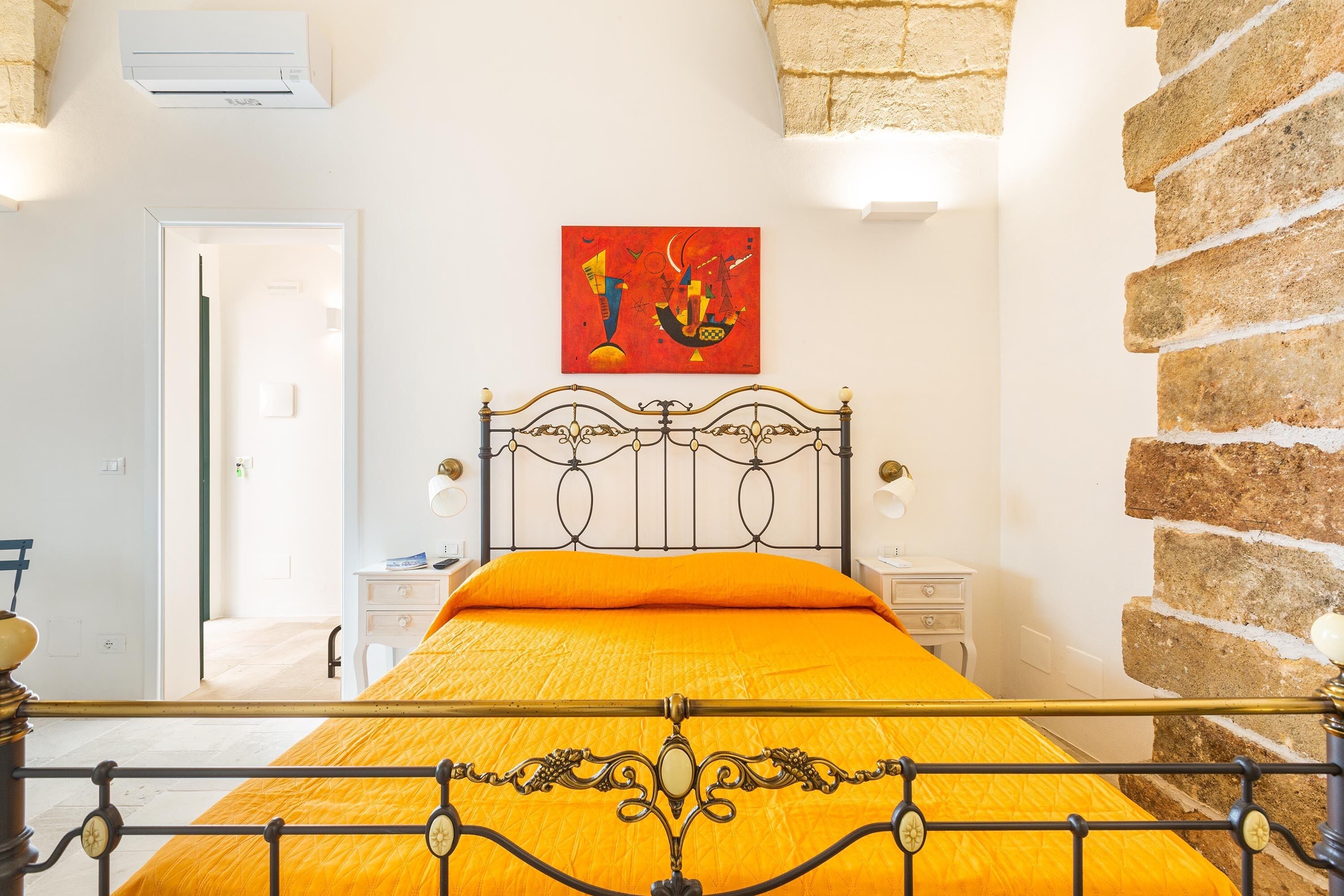 A bedroom with a metal bed frame, a bright orange bedspread, a red abstract painting on the wall, and a brick wall on one side.
