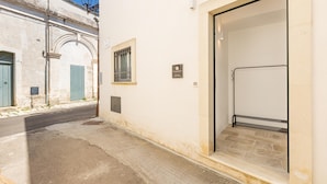 A narrow street with a white building featuring a metal gate, a window with bars, and a door with a sign. - 3398 Leucadense - Grotta 3 by Barbarhouse (Castrignano del Capo)