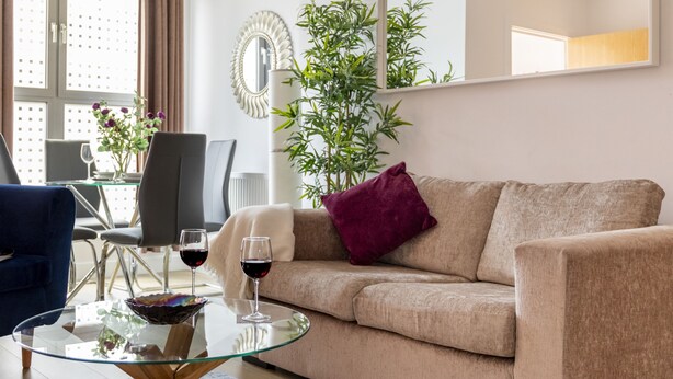 Een woonkamer met een beige bank, een glazen salontafel, twee wijnglazen, een eettafel met stoelen en een grote spiegel.