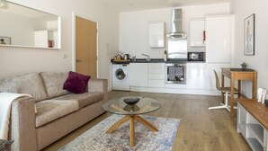 Een moderne woonkamer met een glazen salontafel, een beige bank en een keuken op de achtergrond met een wasmachine, magnetron en oven.