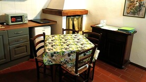 A compact kitchen with a microwave, a small refrigerator, a table with a floral tablecloth, wooden chairs, and a painting on the wall. - Appartamento Olive La Burraia (GAMBASSI TERME)